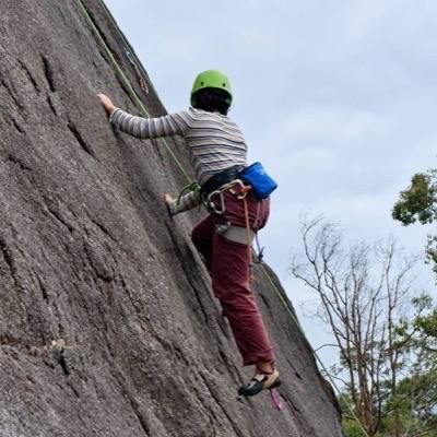 A climber in a green helmet moving up a granite slab, with gum trees behind.
