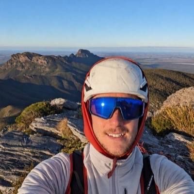 Evan smiling on a mountain ridge in a white helmet and blue sunglasses, peaks behind.