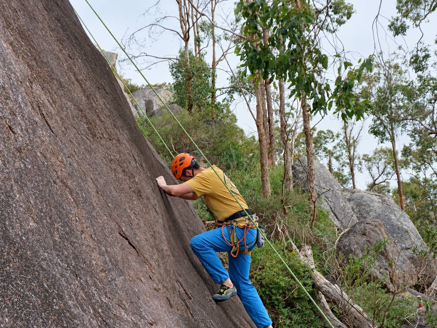 A climber in an orange helmet starting up a granite slab, with gum trees and boulders behind.